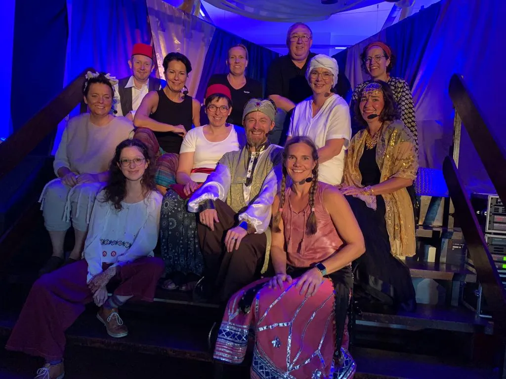 Gruppenfoto des Ensembles von „Der Sultan sucht sein Glück“ in Kostüm vor der Wüstenkulisse mit Palme.
