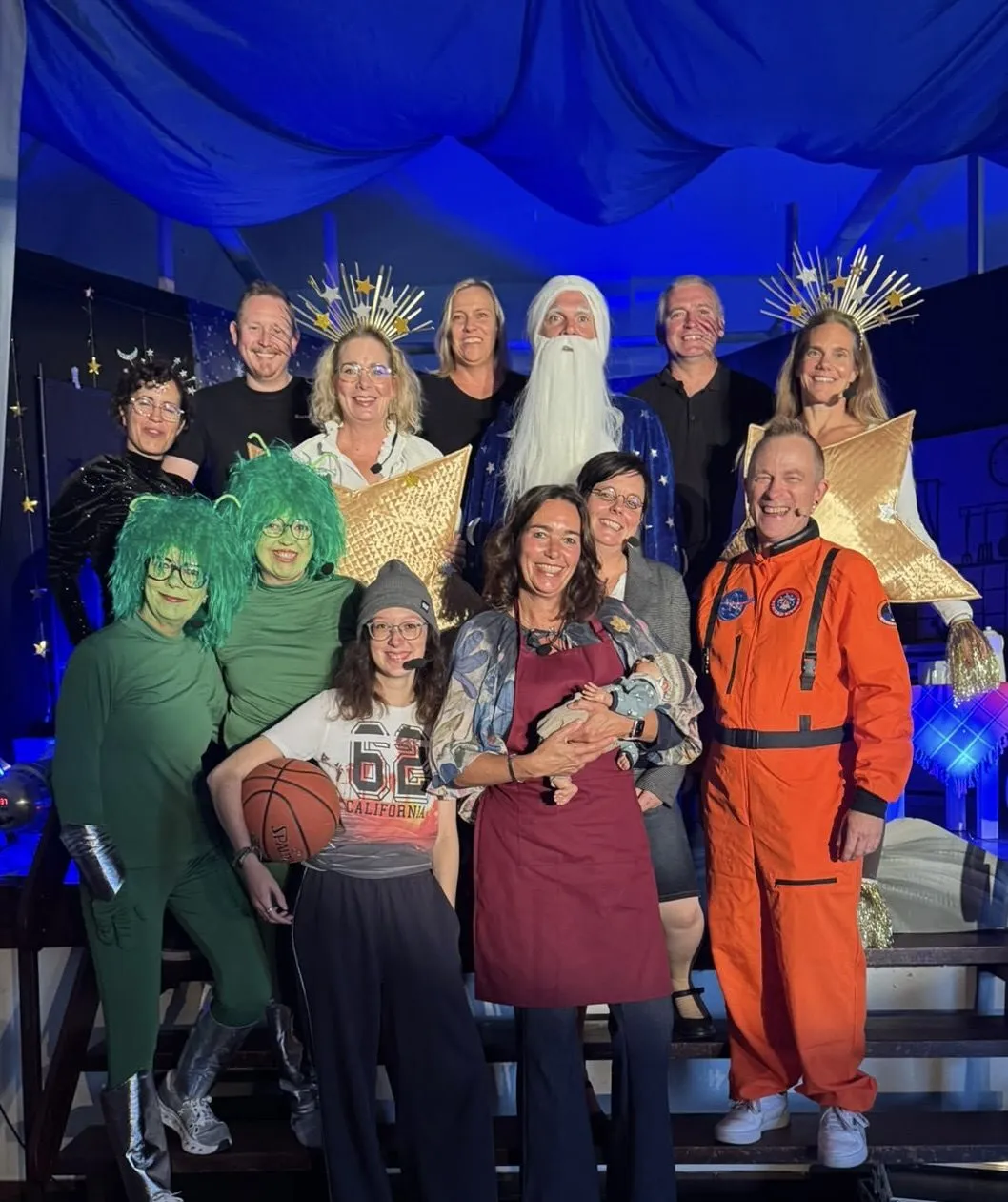 Gruppenfoto des Ensembles von „La Le Lu – hörst du zu?“ in Kostüm — Astronaut, Mann im Mond, Sterne, grüne Außerirdische und weitere Mitwirkende vor blauem Bühnenvorhang.