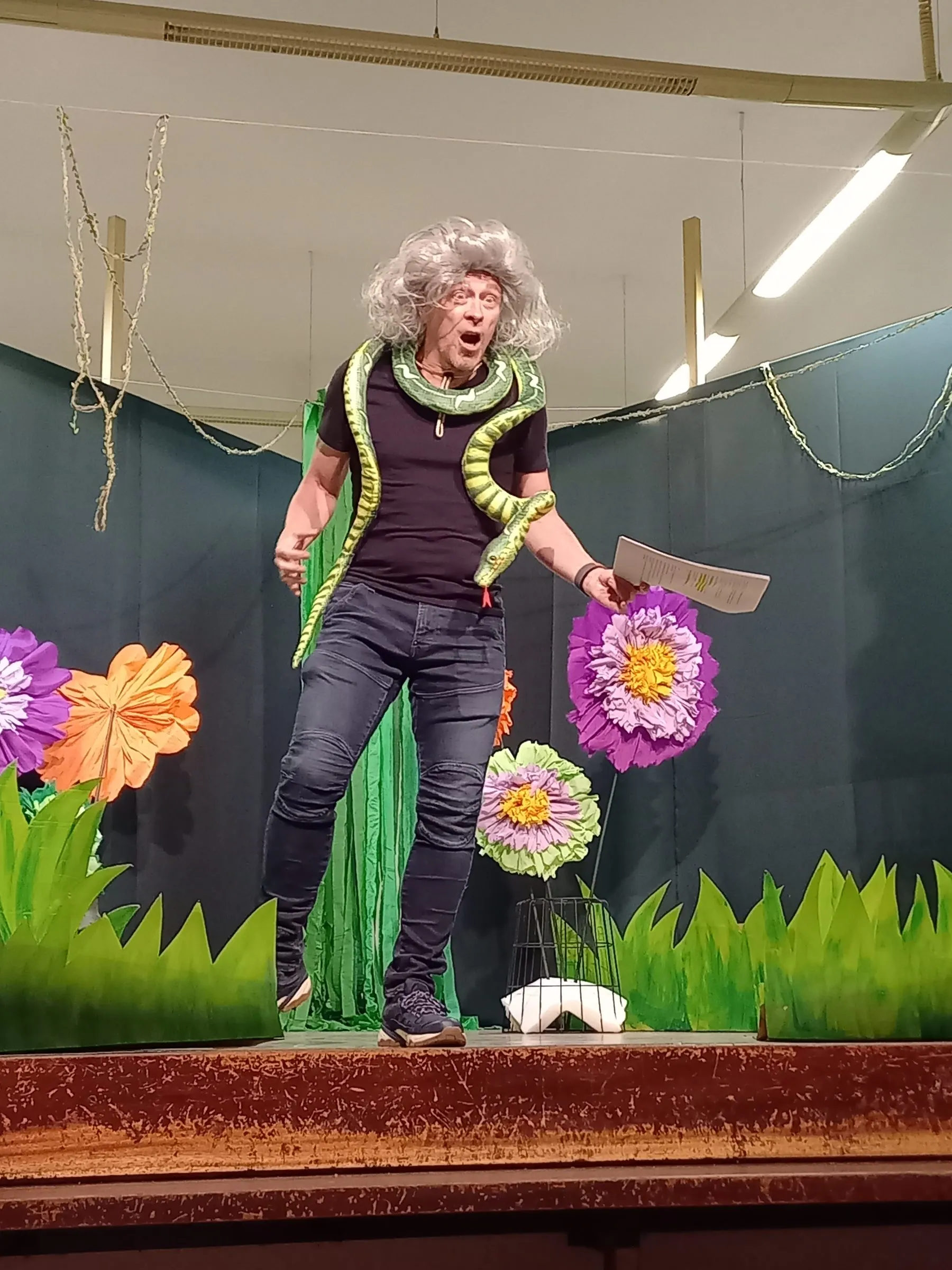 Eine Figur mit grauer Perücke und Schlangenrequisite hält ein Skriptblatt vor der Blumenkulisse.