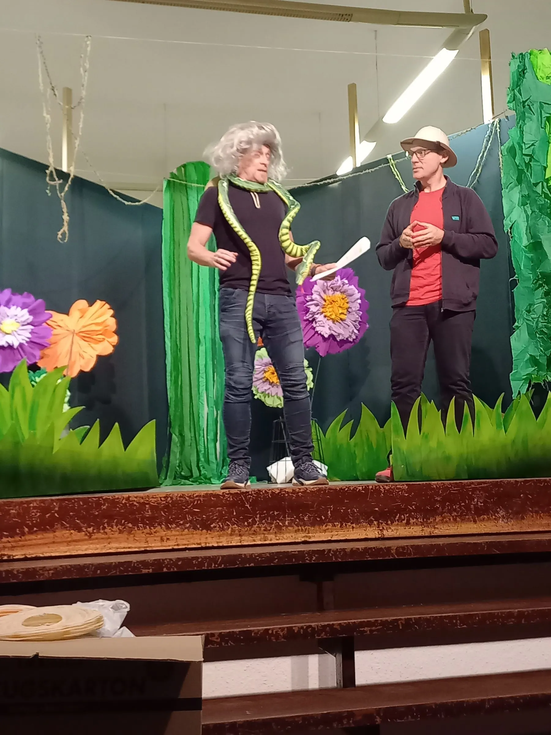 Dieselbe Figur im Gespräch mit einer Forscherin mit Hut vor der Blumenkulisse.