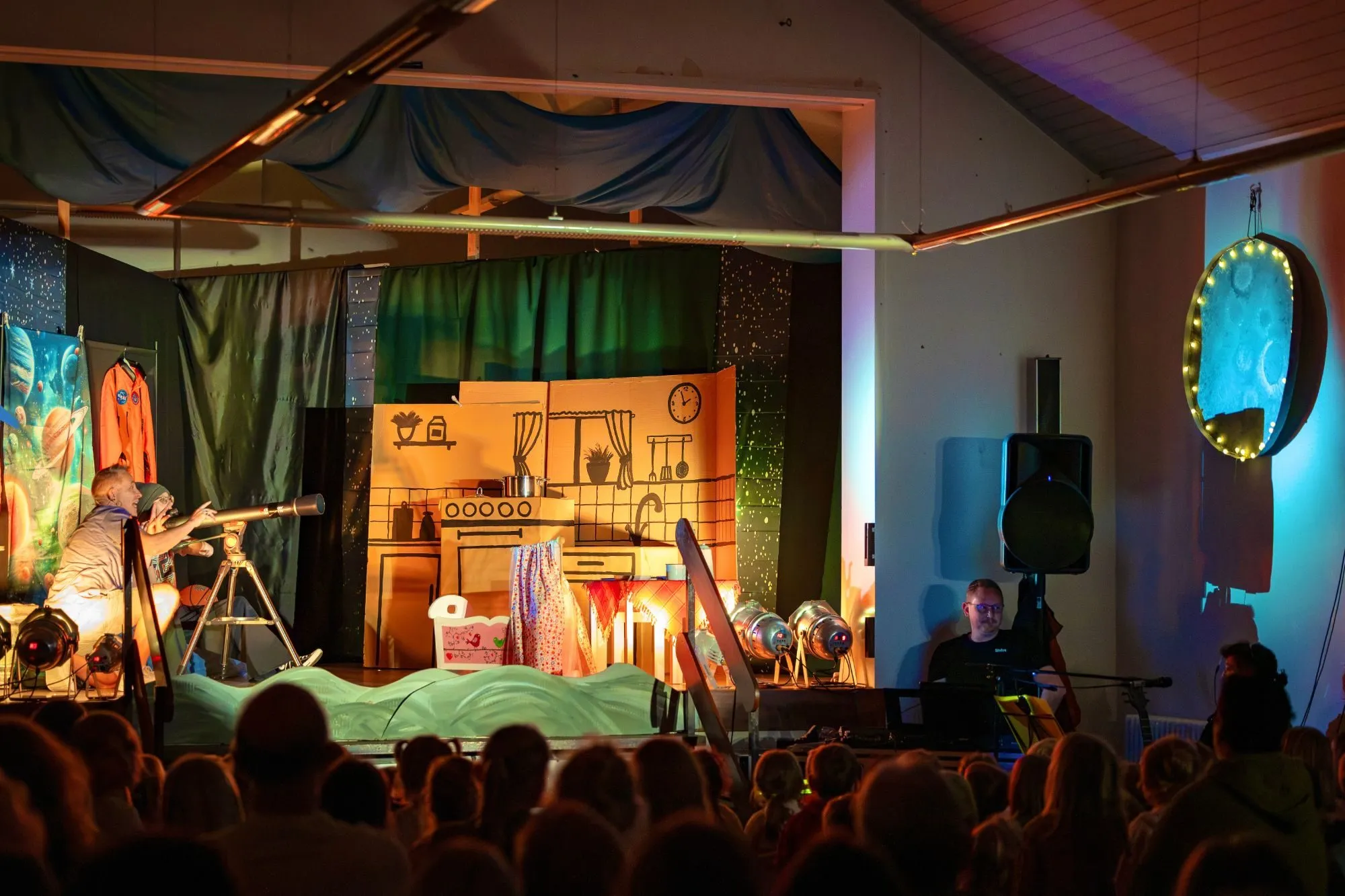 Blick aus dem Publikum auf die Küchenkulisse, links Edwin und Clara am Teleskop, rechts der Musiker am Keyboard und der leuchtende Mond.