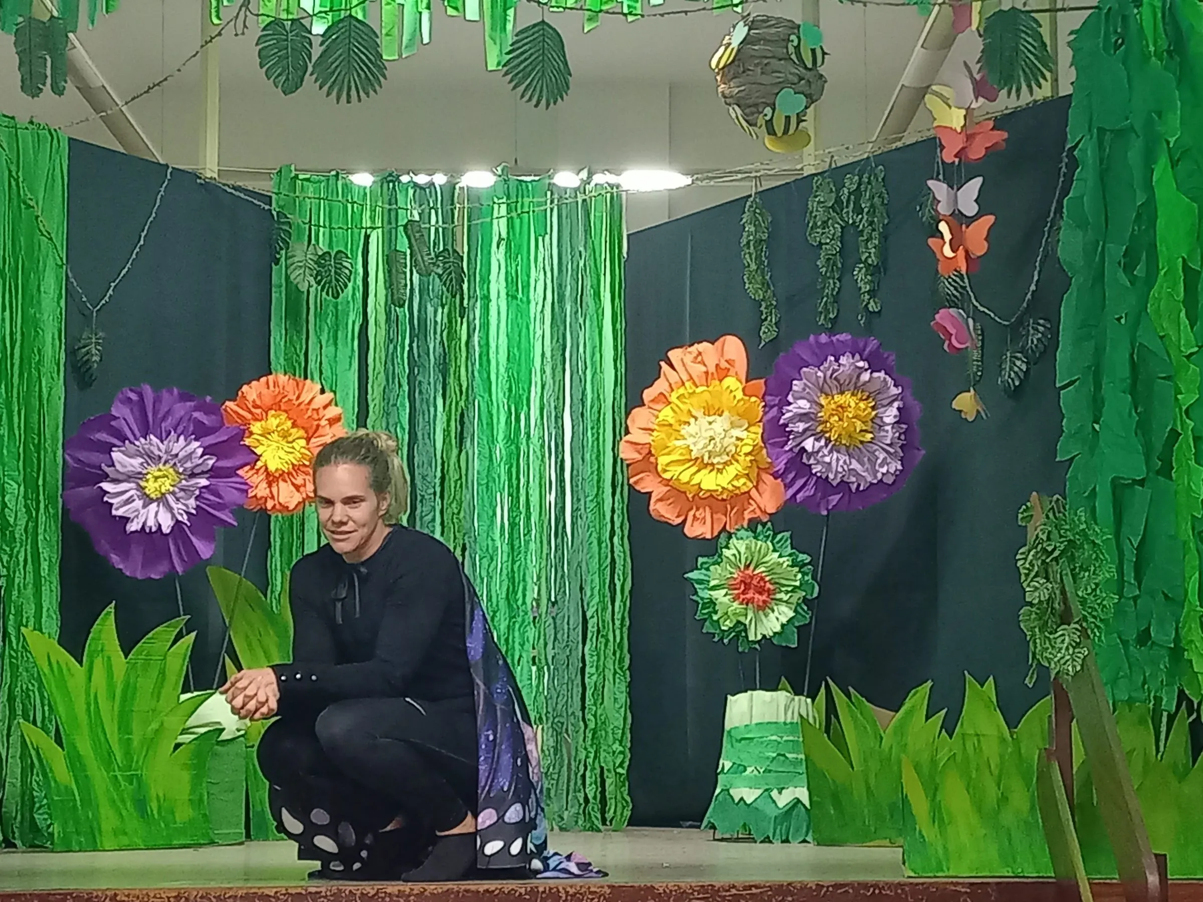 Eine Figur in Schwarz kauert lächelnd vor der Blumenkulisse.